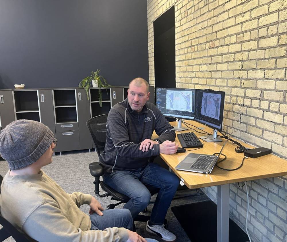 A photo of two people having a mentoring conversation at a workstation with a laptop and two screens.